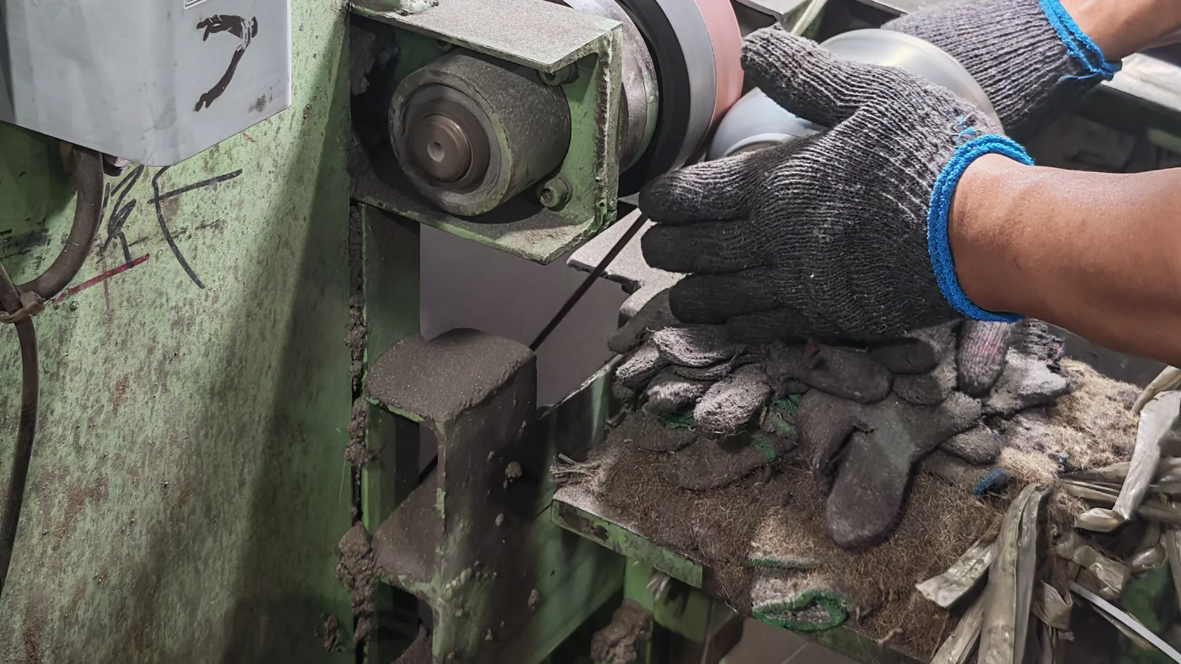 An operator in work gloves hand-grinding a stainless steel body against an abrasive belt on a polishing machine
