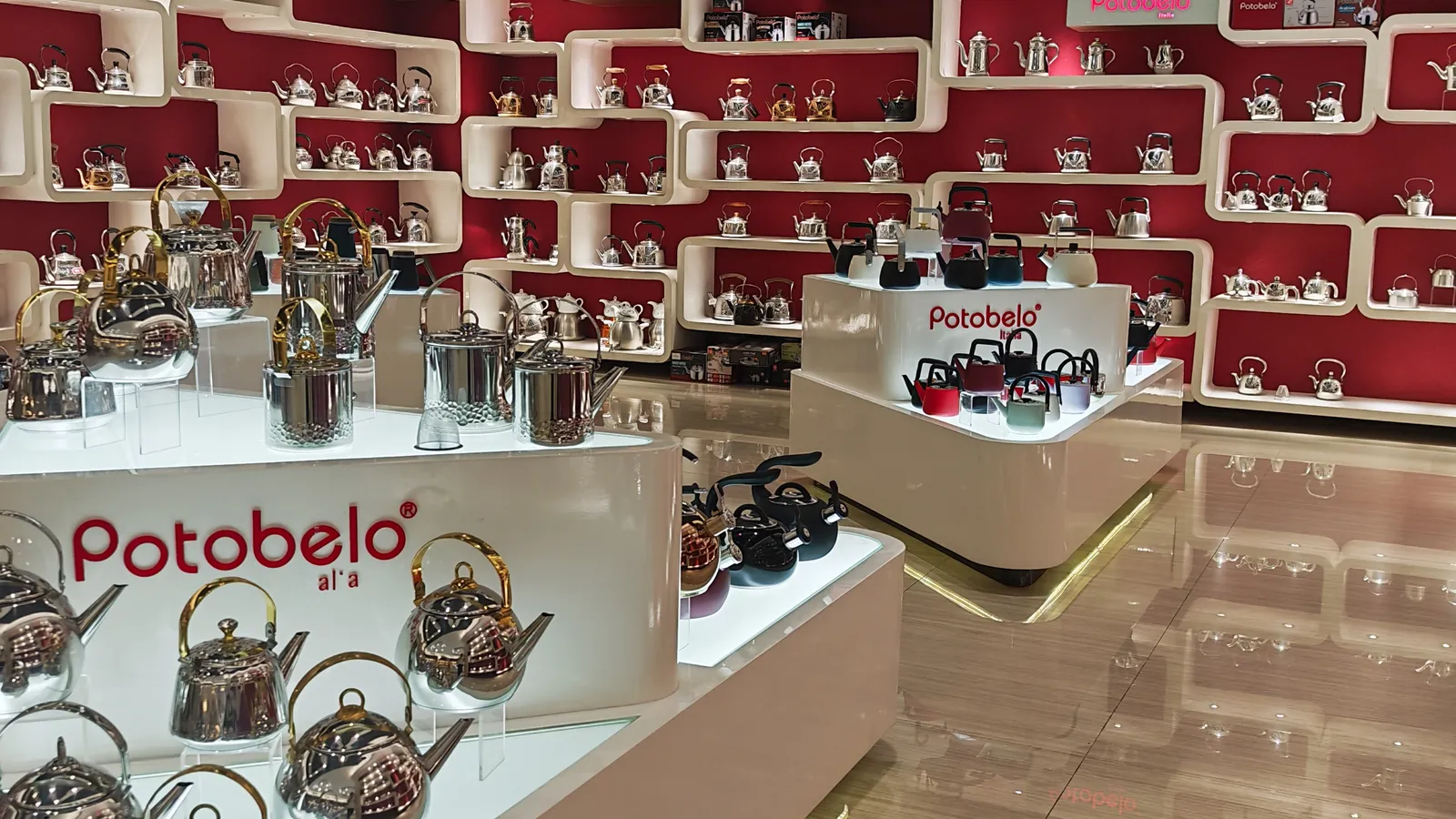 Rows of stainless steel kettles on white showroom shelving against a red wall in the Potobelo showroom