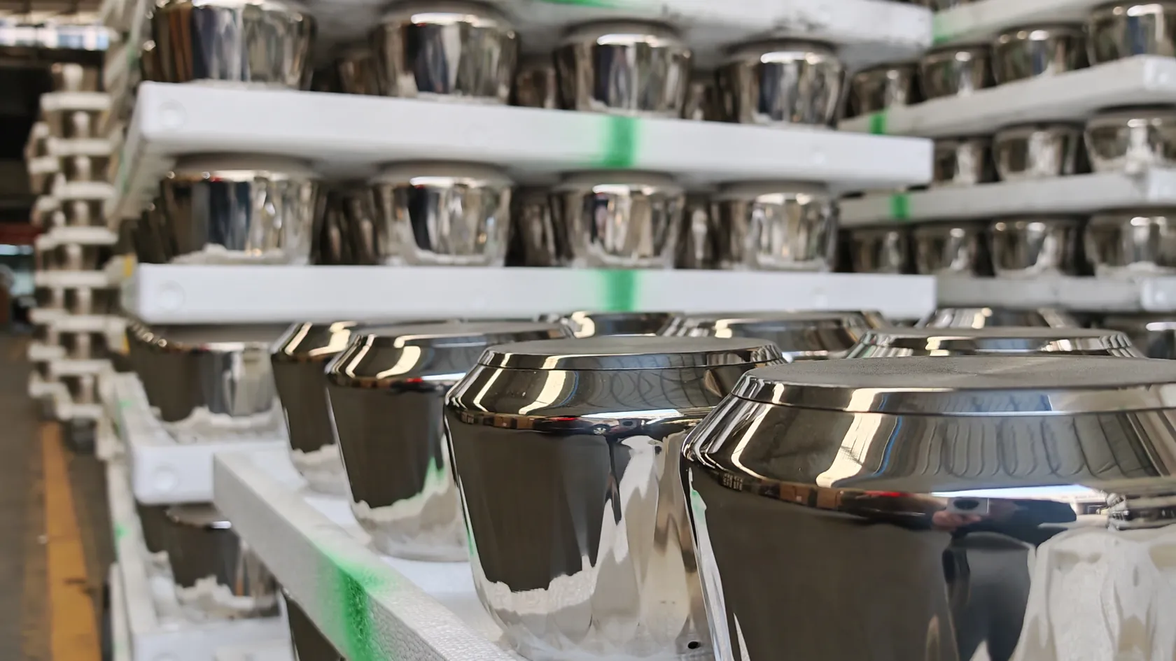 Rows of mirror-polished stainless steel cookware bodies stacked on white racking in a cookware factory