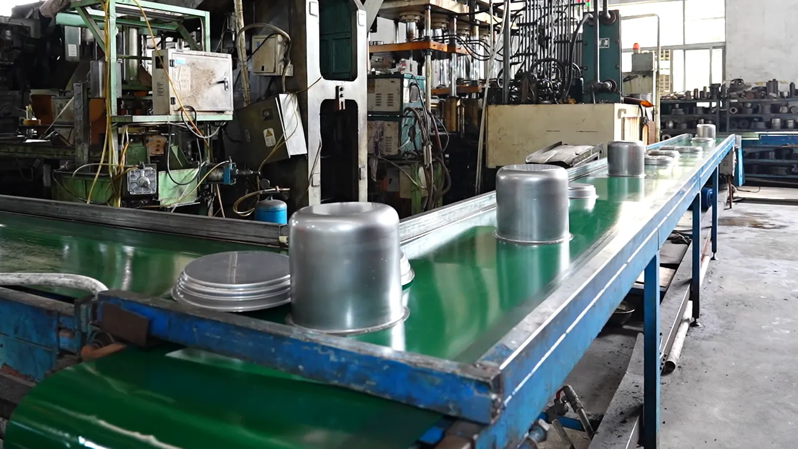 Drawn stainless steel cookware bodies moving along a green conveyor in a stamping shop, with stacked round blanks in the foreground