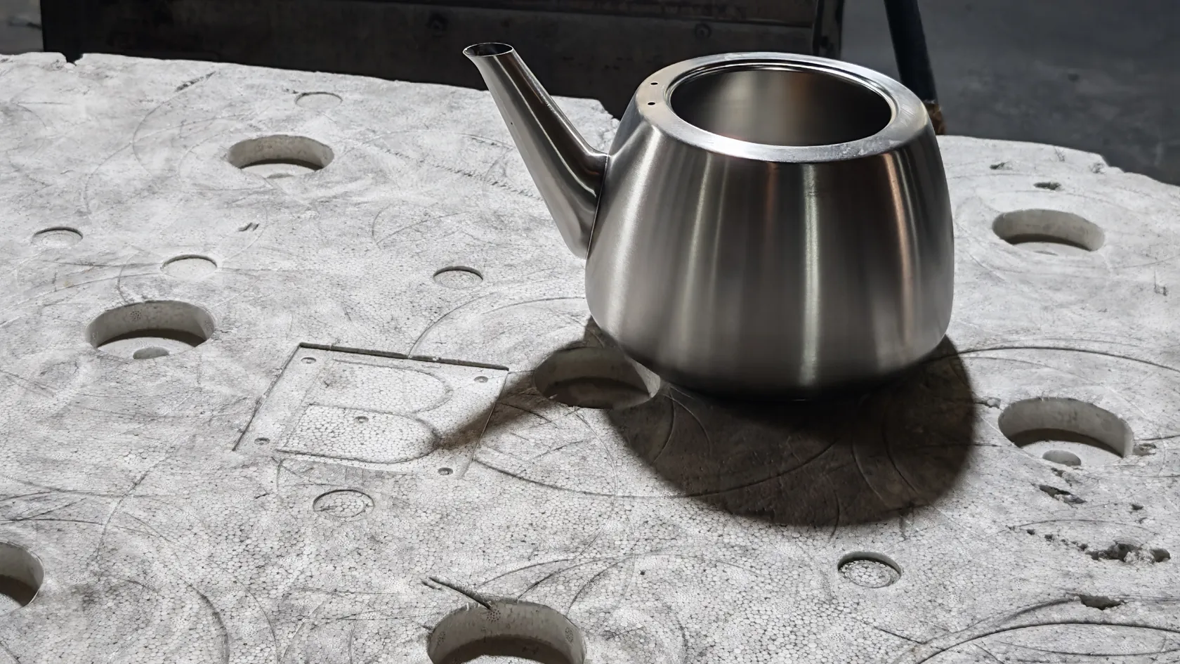 A stainless steel kettle body with its spout, brushed and formed, resting on a perforated work table at the finishing station