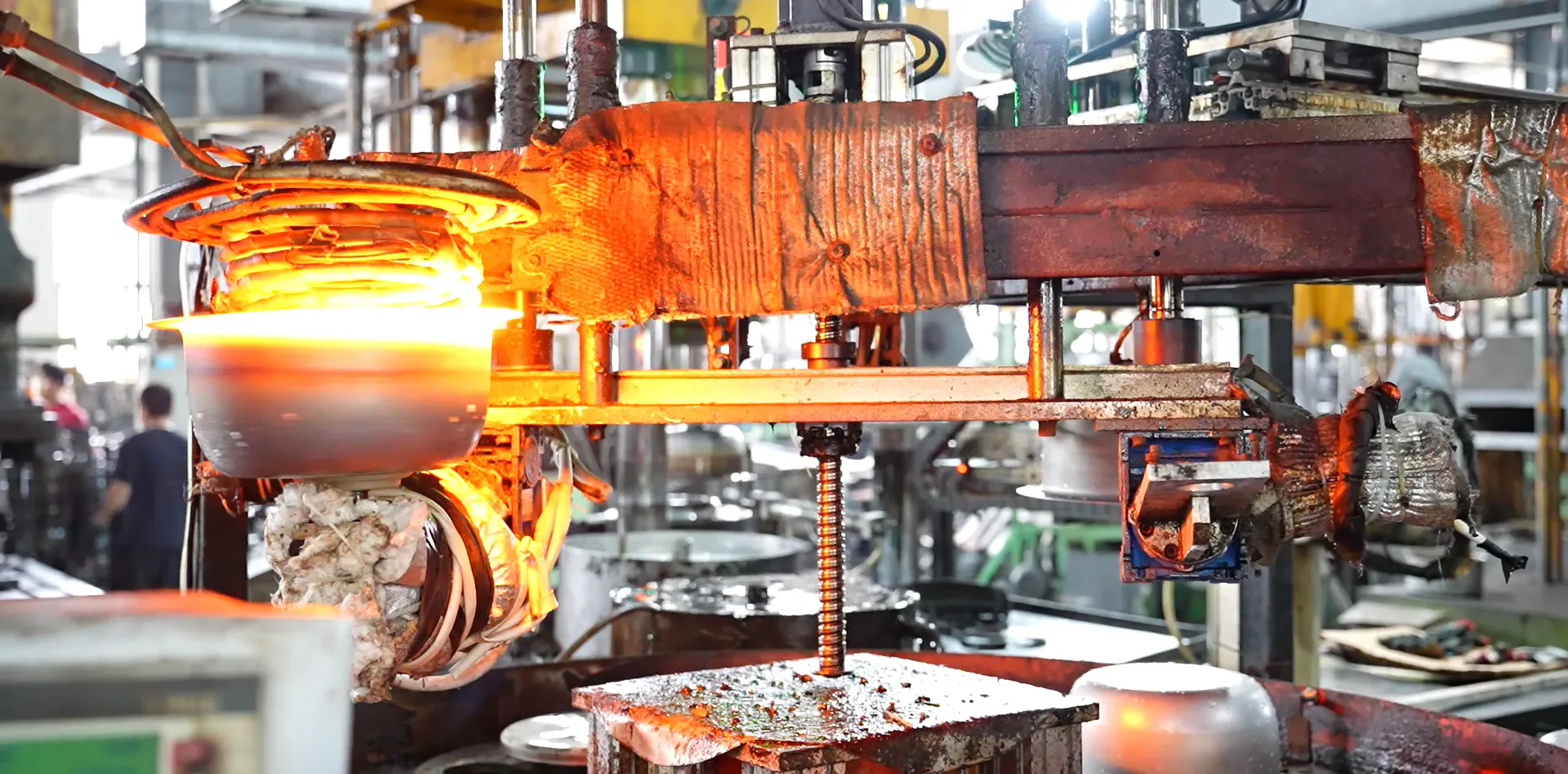 Induction heating station in a cookware factory with a stainless steel workpiece glowing orange inside the coil