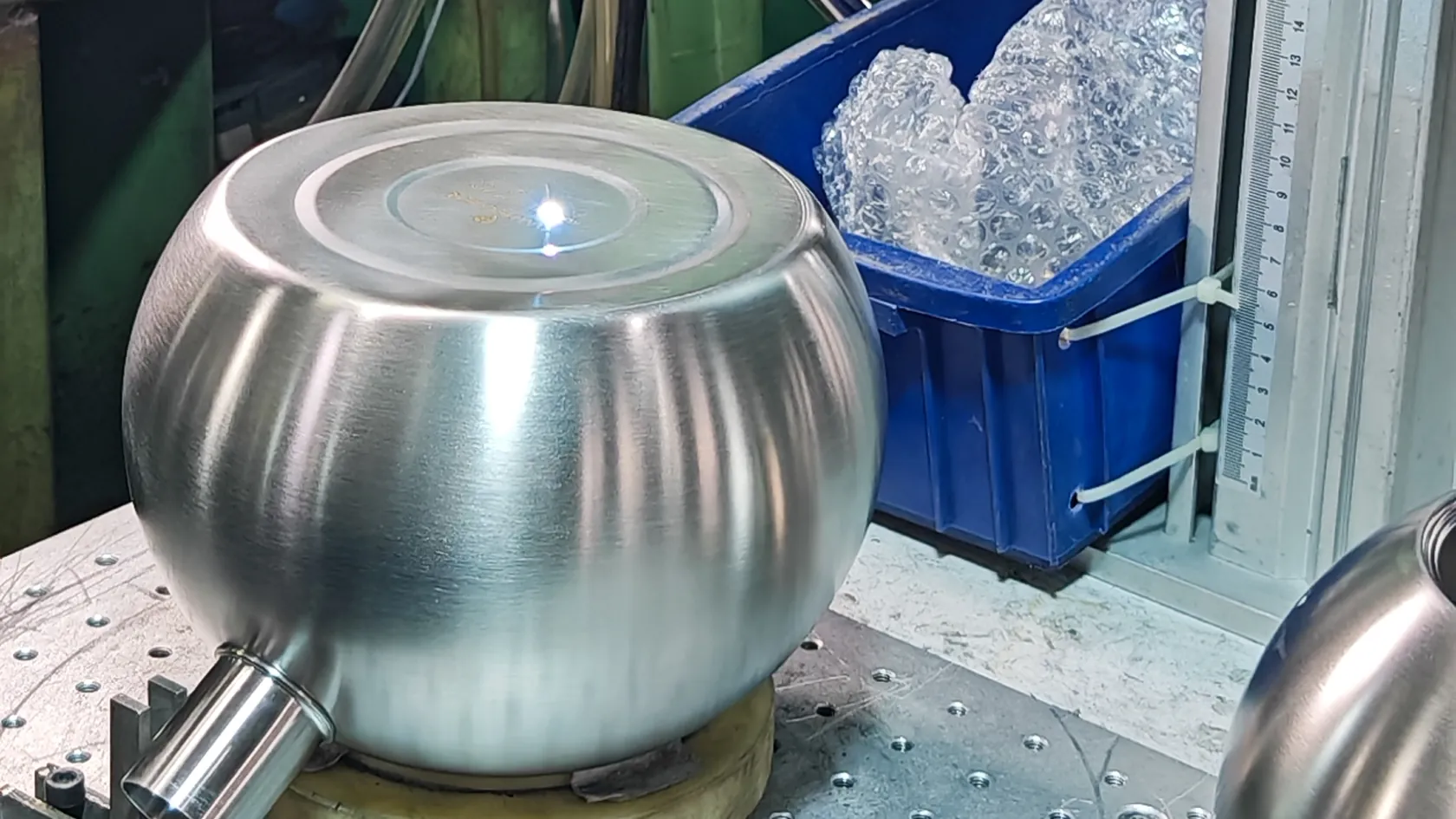 A brushed stainless steel kettle body clamped in a jig at a forming station, with a measuring scale at the edge of the frame