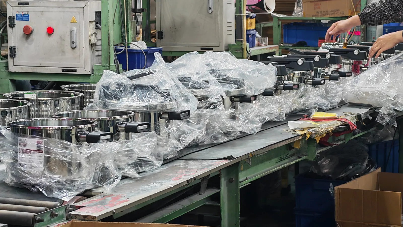 Bagged stainless steel pressure cooker bodies on a packing bench beside an open carton, with a worker's hands at the frame edge