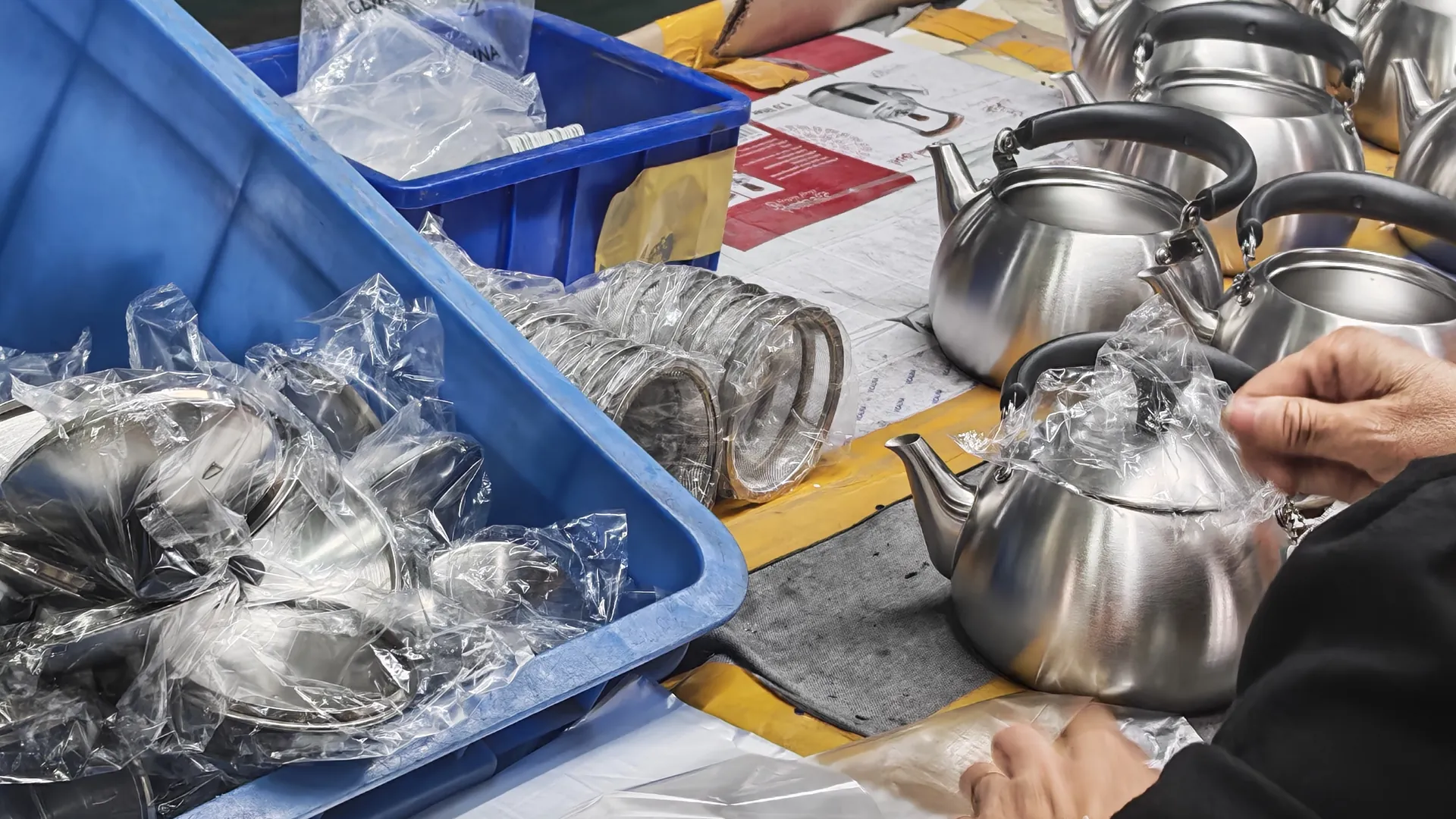 Worker bagging finished stainless steel kettles beside a crate of separately bagged lids and strainers at a packing bench