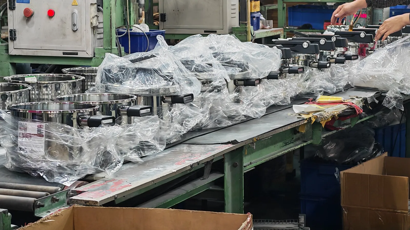 Finished stainless steel pots in poly bags on a packing bench beside cardboard cartons in a cookware workshop