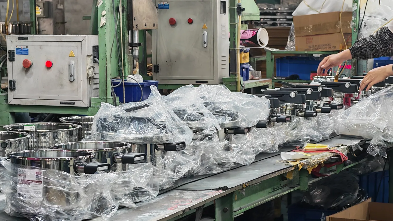 Finished stainless steel pots being packed on a workbench beside stacked cartons in a cookware packing area